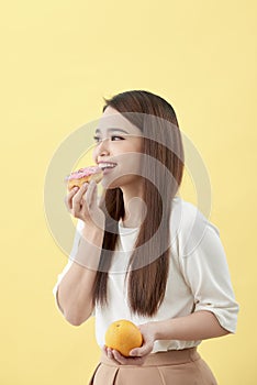 Dieting concept. Beautiful Young Asian Woman holding orange and donut over yellow background. Choosing between junk food with