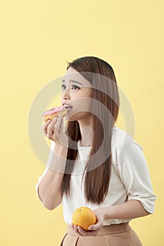 Dieting concept. Beautiful Young Asian Woman holding orange and donut over yellow background. Choosing between junk food with