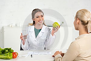 Dietitian in white coat holding apple and glass of water and patient at table