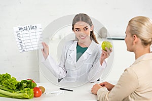 Dietitian in white coat holding meal plan and apple and patient at table