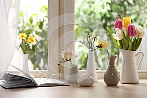 Different beautiful spring flowers, burning candle and book on window sill