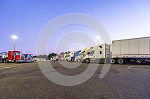 Different big rig semi trucks with semi trailers standing in row on truck stop parking lot at evening time for rest of drivers and