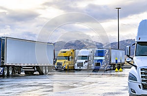 Different big rigs semi trucks with loaded semi trailers standing on the truck stop parking lot with wet surface and mountains on