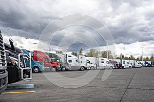 Different big rigs semi trucks with semi trailers standing in long row on truck stop parking lot with stormy sky