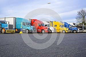 Different big rigs semi trucks with semi trailers standing in row on the reserved spots on truck stop parking lot