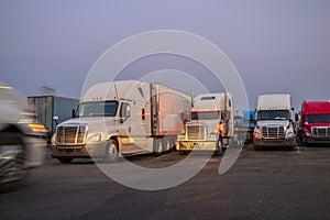 Different big rigs semi trucks with semi trailers standing in row on the twilight truck stop and illuminated by the light of