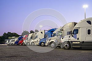 Different big rigs semi trucks standing in row on the night truck stop parking lot with turned on lanterns on poles waiting for
