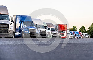 Different Big rigs semi trucks standing in row on truck stop parking lot for rest and comply with the movement schedule