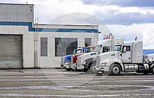 Different big rigs semi trucks tractors standing in row on the warehouse parking lot waiting for next loads for delivery