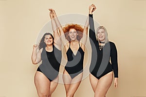 Different Body Types. Group Of Diversity Models Portrait. Smiling Blonde, Brunette And Redhead In Black Bodysuits Holding Hands Up