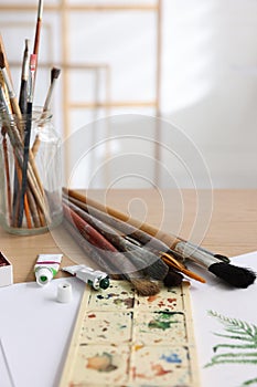 Different brushes, colorful paints, palette and drawing on table in studio. Artist`s workplace