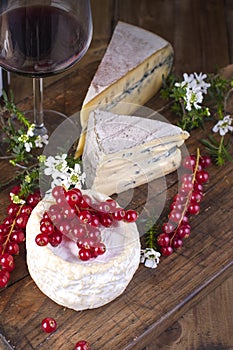 Different cheese with white and blue mold. A glass of red wine and fresh red currant berries. White flowers. Wooden background and