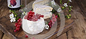 Different cheese with white and blue mold. A glass of red wine and fresh red currant berries. White flowers. Wooden background and