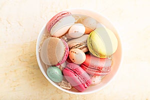 Different colors macaroons and chocolate eggs in ceramic bowl on beige concrete background. top view, close up