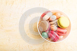 Different colors macaroons and chocolate eggs in ceramic bowl on beige concrete background. top view, copy space