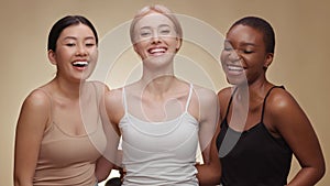 Different female beauty. Three happy diverse young women embracing together and laughing to camera, beige background