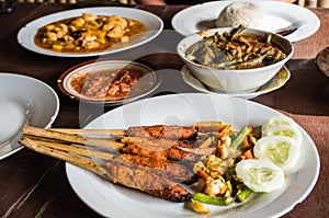 Different Indonesian dishes: Sate Pusut focused foreground, Ikan asam manis, olah-olah, sambal and rice on table