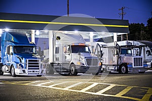 Different make and models big rig semi trucks with semi trailers standing on the truck stop parking lot under the lighted shelter