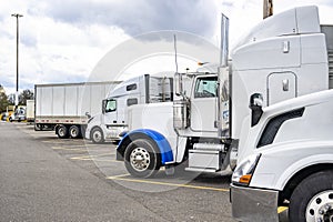 Different make and models of white big rig semi trucks with semi trailers standing in row on truck stop parking lot for truck