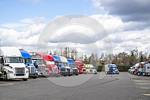 Different makes and models of the big rig semi trucks and semi trailers standing in row on the long truck stop parking lot at
