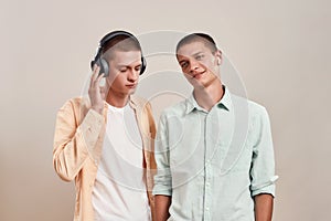 Two young caucasian twin brothers in casual wear listening music in headphones and wireless earphones, while standing