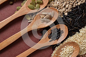 Different seeds in wooden spoons on a surface of different seeds and brown background