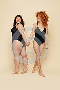 Different Size. Diversity Women Full-Length Portrait. Smiling Brunette And Redhead In Black Bodysuits Posing On Beige Background. photo