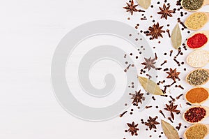 Different spices in bamboo spoons and scattered spices on white wood background, top view, closeup.