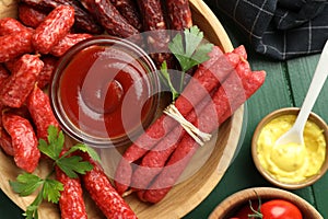 Different thin dry smoked sausages, parsley and sauces on green wooden table, flat lay