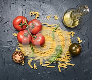 Different types of pasta, laid out with various ingredients, tomatoes, herbs, oil in a decanter and seasonings on a dark rustic ba