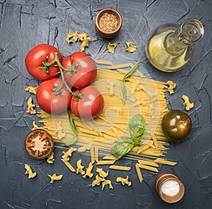 Different types of pasta, laid out with various ingredients, tomatoes, herbs, oil in a decanter seasonings on a dark rustic ba