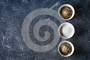 Different types of salt in glass bowls on a dark gray table.