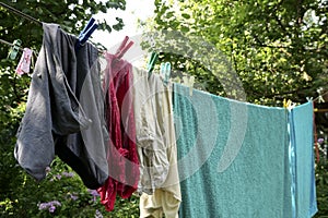 Different washed clothes is dried on rope in backyard or garden. Drying linen with multicolored clothespins. Summer sunny day.