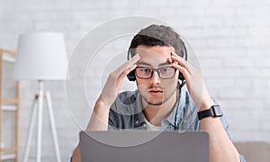 Difficult task. Concentrated guy in glasses and headphones puts hands to forehead and ponders something, looks at laptop