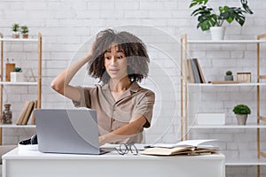 Difficult task or problems at online work. Surprised african american woman scratches her head and looks at laptop