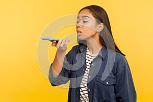 Digital assistant app. Portrait of girl in denim shirt talking to mobile phone, using virtual voice service to order online