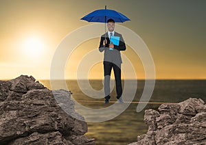 Digital composite image of businessman standing on rope holding diary with blue umbrella amidst rock