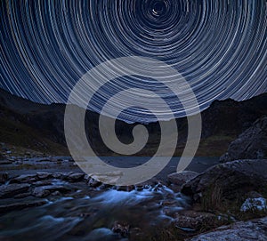 Digital composite image of star trails around Polaris with landscape image of river flowing down mountain range near Llyn Ogwen