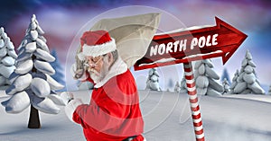 North Pole text and Santa holding sack with Wooden signpost in Christmas Winter landscape