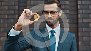 Digital currency. Handsome businessman in formal wear and eyeglasses holding bitcoin in one hand and looking at it while