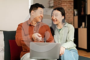 Digital entertainment. Excited asian couple using laptop at home, relaxing in living room with computer, watching movies