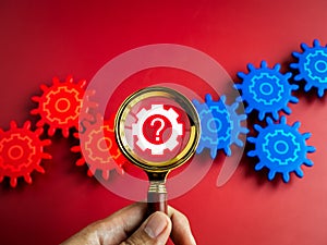 Digital gear icon wheel with question mark symbols on magnifying glass lens in hand while looking through on red gears on red.