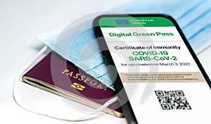 The digital green pass of the european union with the QR code on the screen of a mobile phone over a surgical mask and a passport