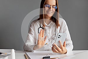 Digital health specialist. Smiling woman doctor wearing white medical lab coat stethoscope and glasses using smart phone for