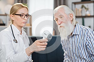 Digital infrared thermometer in hand of pleasant blond 40-aged woman doctor or nurse, visiting her senior male patient