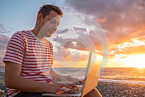 Digital nomad man working with laptop sits on the beach by the sea during sunset. Male freelancer work online from a