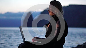 Digital Nomad, a young tattooed man working remotely online, typing on a laptop keyboard while sitting on a beach at