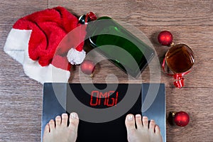 Digital scales with female feet on them and sign omg! surrounded by Christmas decorations, bottle and glass of alcohol.