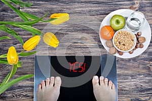 Digital scales with female feet on them and sign yes! surrounded by spring flowers, plate with healthy food and glass of water.