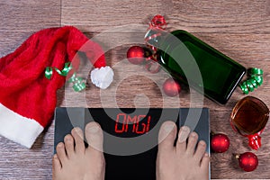 Digital scales with male feet on them and sign omgsurrounded by Christmas decorations, bottle and glass of alcohl.